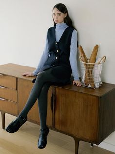 Woman in a blue turtleneck and dark vest dress sitting on a wooden cabinet