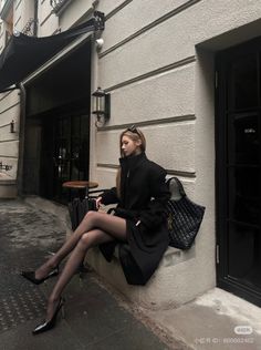 Woman in a black coat and tights sitting on a ledge outdoors