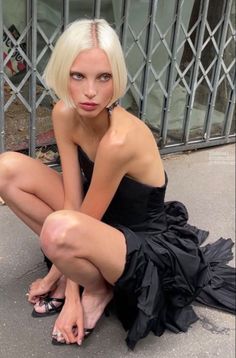 Woman with blonde bob hair in a black strapless dress squatting outdoors