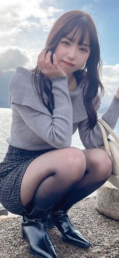 Woman in a grey sweater and mini skirt squatting outdoors by the water