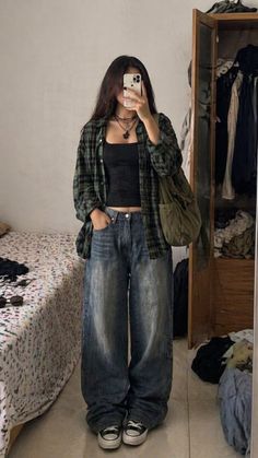 Person in a plaid shirt and baggy jeans taking a mirror selfie indoors