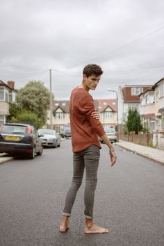 Young man in a rust-colored shirt and grey jeans standing barefoot on a street