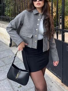 Woman in a gray jacket and black mini skirt holding a black handbag outdoors