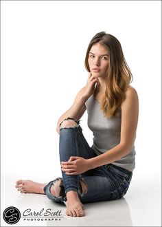 Woman with long hair wearing ripped jeans and a gray tank top, sitting barefoot