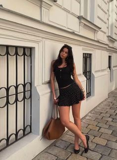 Woman in a black tank top and polka dot skirt leaning against a white building