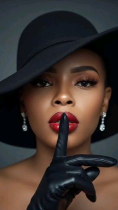 Woman in a black hat and gloves with red lipstick, making a 'shush' gesture
