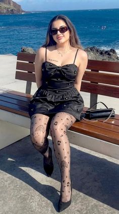 Woman in a black dress and patterned tights sitting on a bench by the ocean