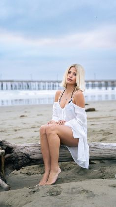 Woman with blonde hair sitting on driftwood on a beach with a pier in the background