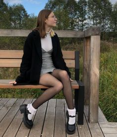 Woman in a black coat and gingham dress sitting on a wooden bench outdoors