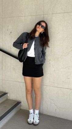 Woman in a gray cardigan and black mini skirt leaning against a wall