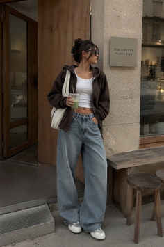 Woman in a brown hoodie and wide leg jeans holding a drink outside a coffee shop