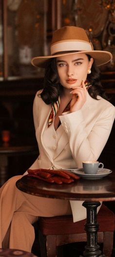 Woman in a fedora hat and white suit sitting at a table with a coffee cup