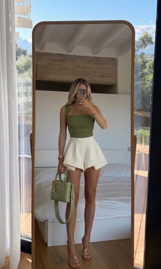 Woman in a green top and white shorts taking a mirror selfie in a bedroom