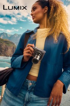 Woman with curly hair holding a camera outdoors with mountains and ocean in the background