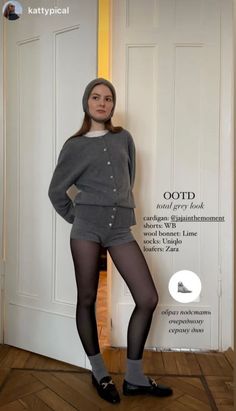 Woman in a grey cardigan, shorts, and black tights standing indoors