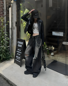 Person in a black leather jacket, ripped jeans, and baseball cap standing outdoors