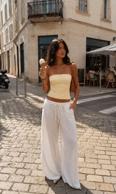 Woman in a yellow strapless top and white wide-leg pants holding an ice cream cone on a cobblestone street