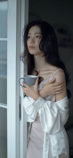 Woman with long dark hair holding a mug, wearing a white shirt over a slip dress