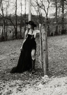Woman in a black dress, cowboy hat, and boots leaning on a fence post in a forest