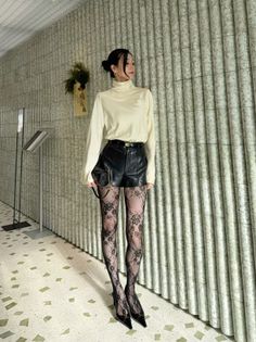 Woman in a cream turtleneck, leather shorts, and lace tights posing indoors