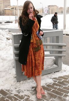 Woman in an orange dress and black coat standing barefoot in the snow