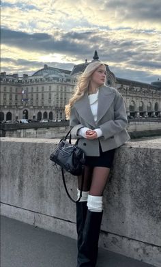 Woman with blonde hair in a grey jacket and black skirt leaning on a stone wall