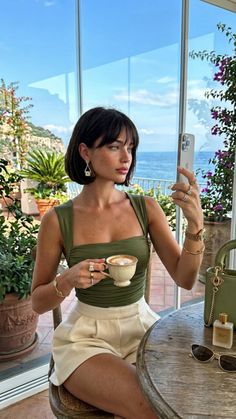 Woman with short hair and a green top holding a coffee cup, taking a selfie