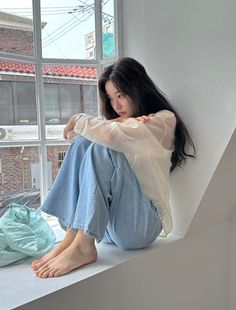 Woman with long dark hair sitting on a window sill in a white sheer top and jeans