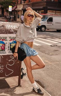 Woman in sunglasses and a crop top leaning against a post on a city street