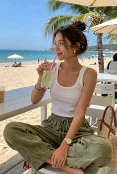 Woman in a white tank top and green pants drinking a beverage at the beach