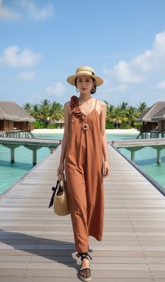 Woman in a brown jumpsuit and straw hat walking on a wooden pier over clear blue water