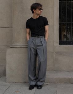 Man in a black t-shirt and grey pants leaning against a stone wall