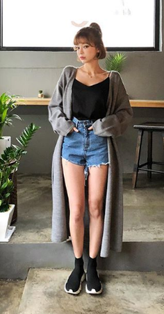 Woman in denim shorts, black top, and long gray cardigan indoors