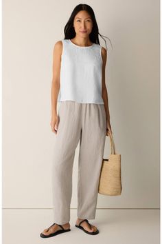Woman in a white sleeveless top and linen pants holding a straw bag