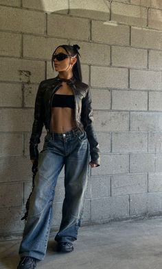 Woman in a leather jacket, crop top, and baggy jeans against a concrete wall