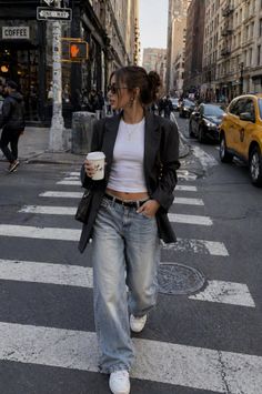 Woman in a blazer and loose jeans walking across a city street with a coffee cup