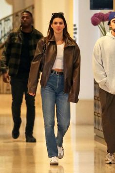 Woman in a brown leather jacket and jeans walking indoors with other people