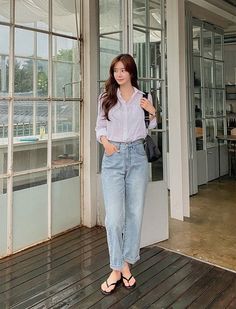 Woman in a white shirt and blue jeans standing indoors by a large window