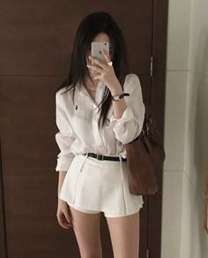 Woman in a white shirt and shorts taking a mirror selfie indoors