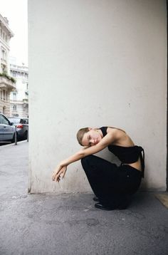 Woman in black top and pants squatting on a street next to a wall