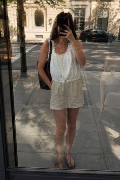 Woman in a white top and linen shorts taking a mirror selfie on a street
