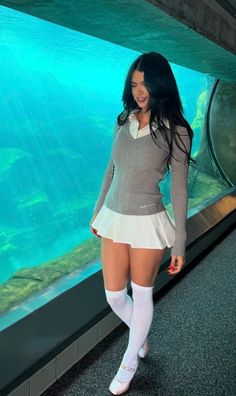 Woman in a white pleated skirt and grey sweater posing in an aquarium