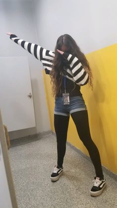 Person in a striped sweater and denim shorts doing a dab pose indoors