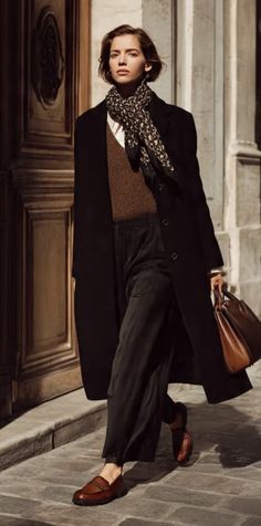 Woman in a black coat and brown sweater walking outdoors with a brown handbag
