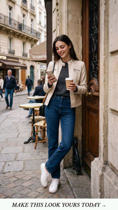 Woman in a leather jacket and jeans holding a phone and coffee on a street