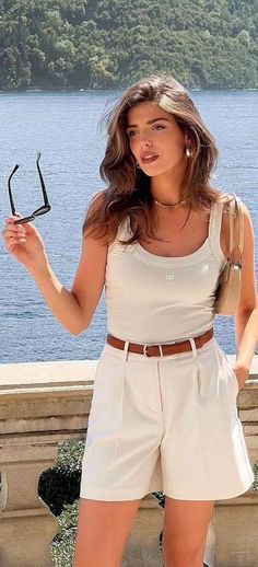 Woman in a white tank top and shorts holding sunglasses by the water