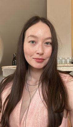 Woman with long brown hair wearing a pink top and layered necklaces indoors