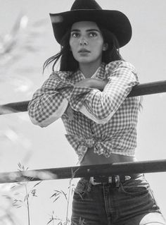Woman in a cowboy hat and gingham shirt leaning on a fence outdoors