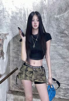 Woman in a black crop top and camouflage skirt holding a phone and blue bag
