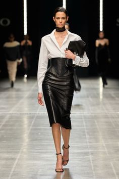 Model in a white shirt, leather corset, and skirt on a runway